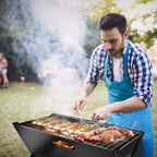 Man wearing a blue apron cooking sausages and meat on a smoking portable charcoal barbecue grill outdoors
